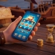 An illustration of a hand holding a phone with a pirate-themed slot game on screen, beside a small stack of coins and a sand timer — learning a Jili game with a small, time-limited first session.